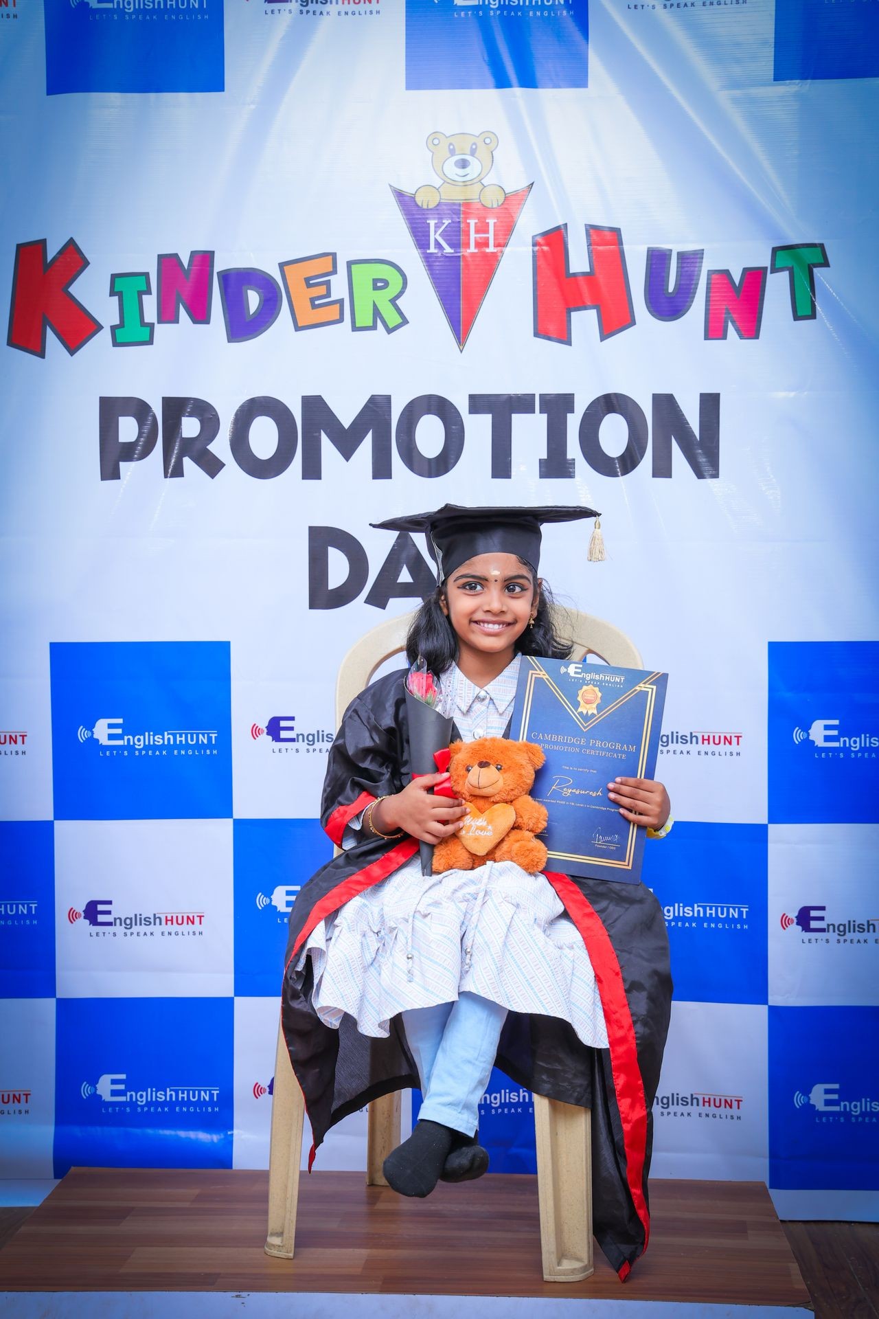 Child in graduation gown holding certificate and teddy bear at promotion day event.