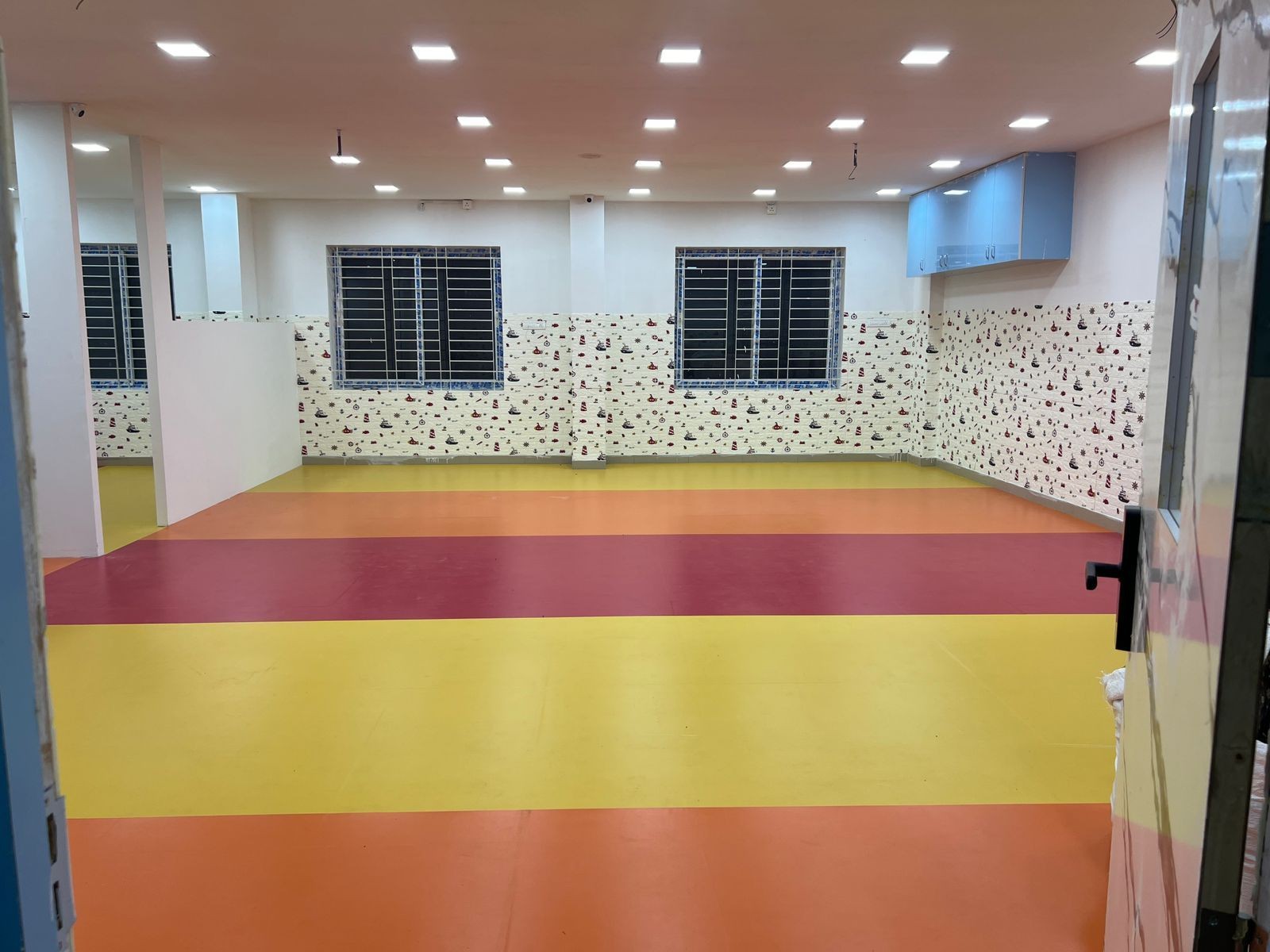 Empty room with colorful striped floor and patterned wallpaper, featuring large windows and ceiling lights.