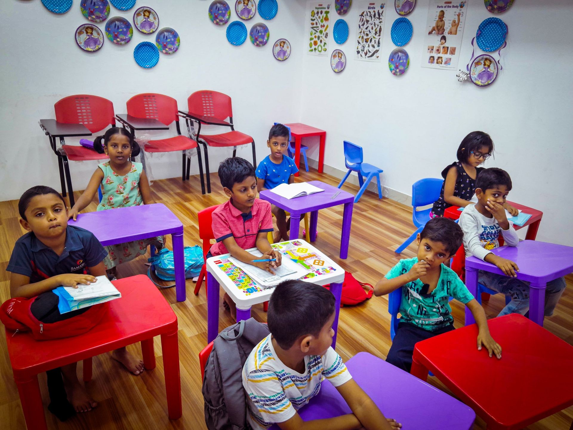 Children sitting at colorful desks in a classroom with educational wall decorations and hardwood flooring.