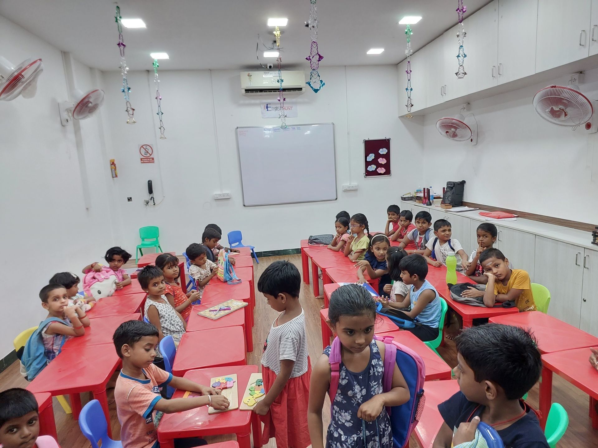 Children in a classroom sitting at red tables with toys and school supplies, decorated with hanging ornaments.