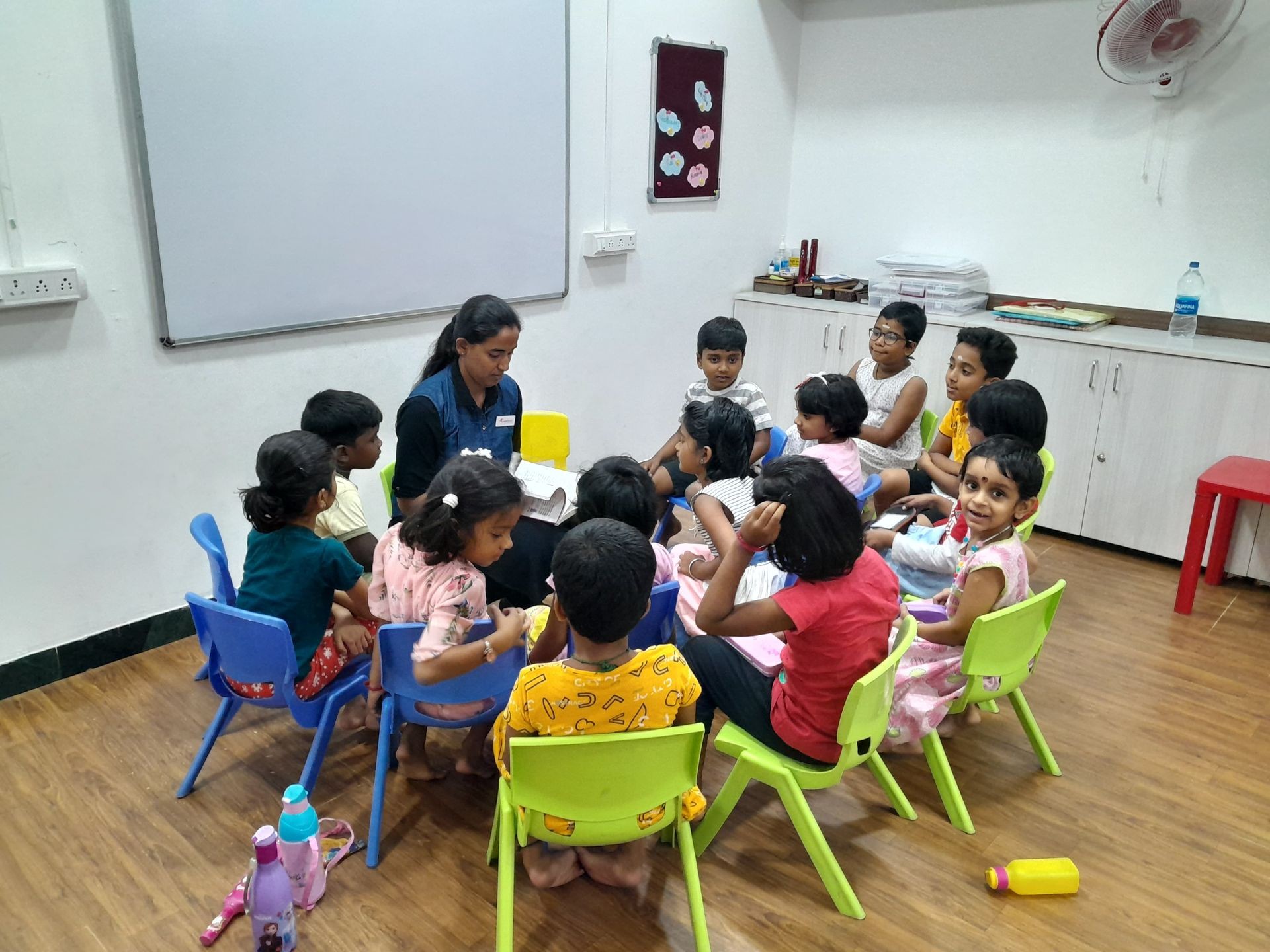 Teacher reading a book to a group of young children seated on colorful chairs in a classroom.