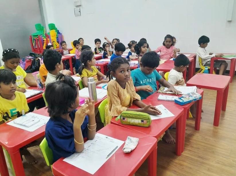 Group of children sitting at red desks in a classroom, engaged in writing activities.