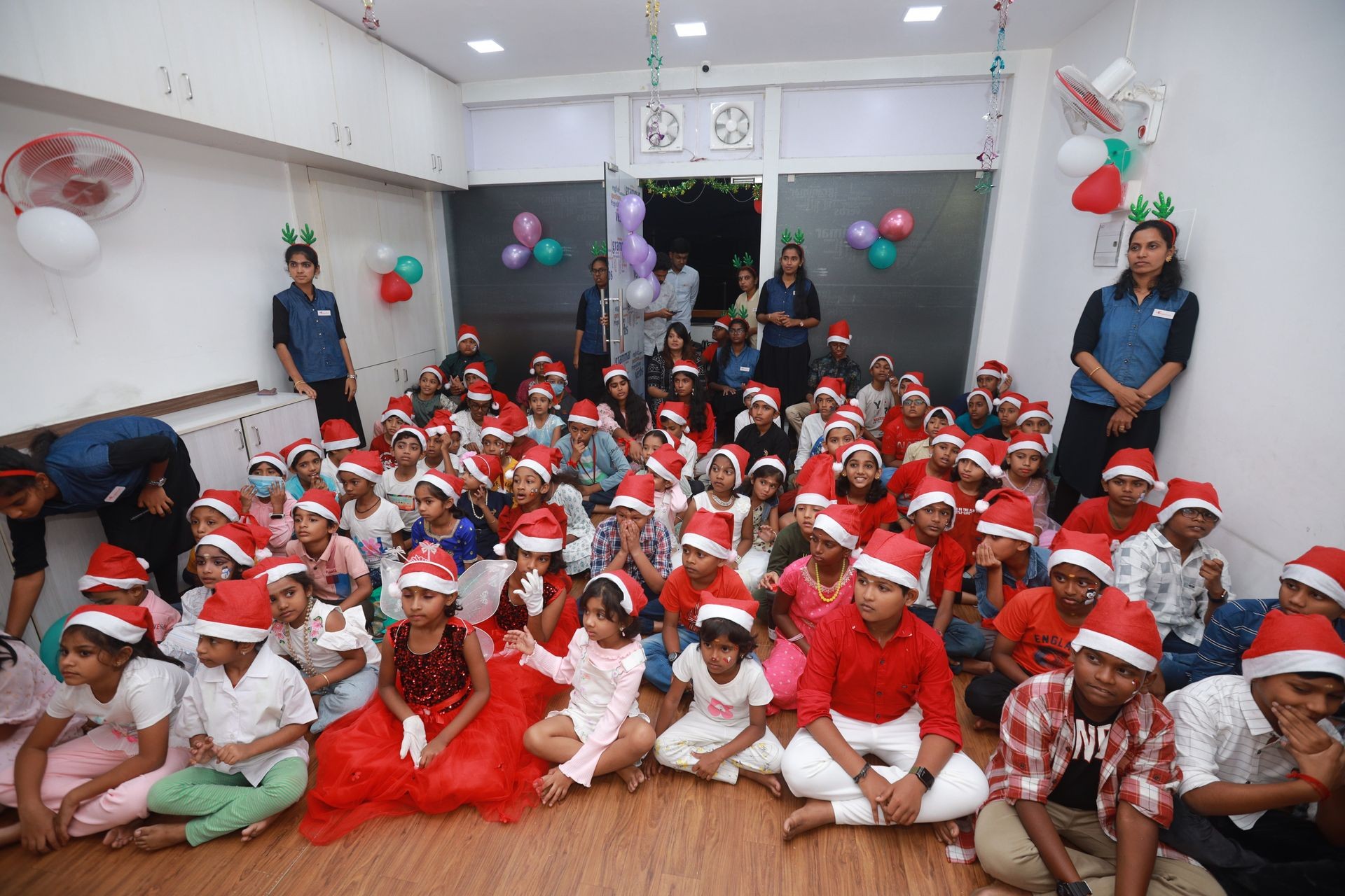 Group of children and staff wearing Santa hats seated in a festive room with balloons and holiday decorations.